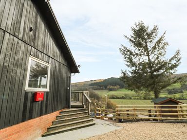 An exterior view of a building with wooden siding steps and a railing overlooking a rural landscape with a tree and fenced area at Middle Hivron in Bleddfa