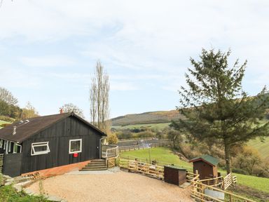 A black house with a gravel driveway near fenced grassy hills and trees at Middle Hivron in Bleddfa