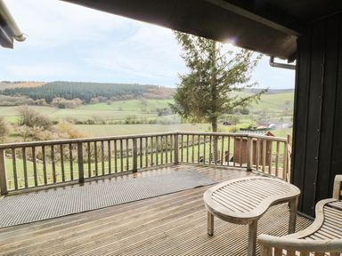 A wooden deck with a railing a curved wooden bench and table overlooking fields and hills at Middle Hivron in Bleddfa