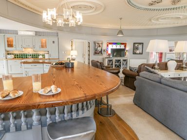 An open plan living room and kitchen with a wooden breakfast bar croissants and juice on the counter and sofas with lamps at Capel Mawr in Llanrug near Llanberis