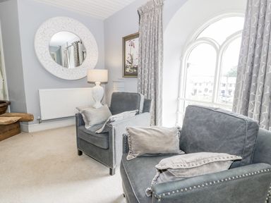 Two gray armchairs with pillows near an arched window with curtains and a round decorative mirror on the wall at Capel Mawr in Llanrug near Llanberis