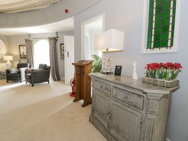 A room with two armchairs near a window with curtains and a wooden cabinet with a lamp and flowers on top at Capel Mawr in Llanrug near Llanberis
