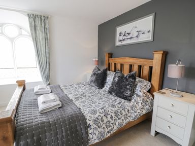 A bedroom with a wooden bed frame a floral bedspread two gray pillows folded towels on the bed a white bedside table with a lamp and a window with patterned curtains at Capel Mawr in Llanrug near Llanberis