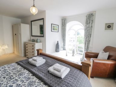 A bedroom with a wooden bed with towels on the bed a dresser with jars an armchair and an arched window with curtains at Capel Mawr in Llanrug near Llanberis