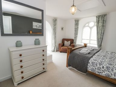 A bedroom with a wooden bed, white chest of drawers, armchair, and arched window at Capel Mawr in Llanrug near Llanberis