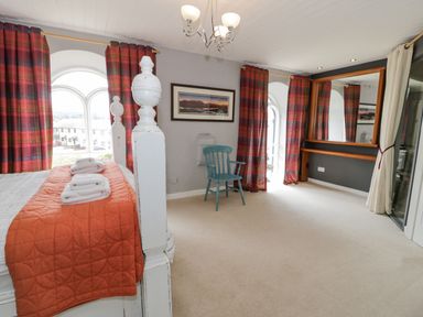 A bedroom with a white bed and orange bedspread and a blue chair near windows with red plaid curtains at Capel Mawr in Llanrug near Llanberis