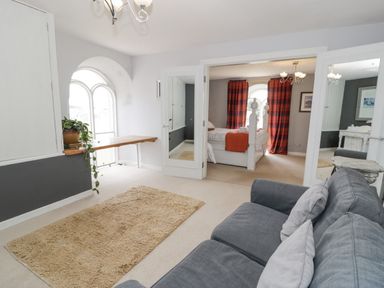 A sitting area with a gray sofa and beige rug connected to a bedroom with a white bed and red plaid curtains at Capel Mawr in Llanrug near Llanberis