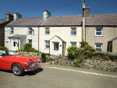 A row of terraced houses with stone walls and a red vintage car on the road outside at Bwthyn Megan in Llangaffo near Newborough