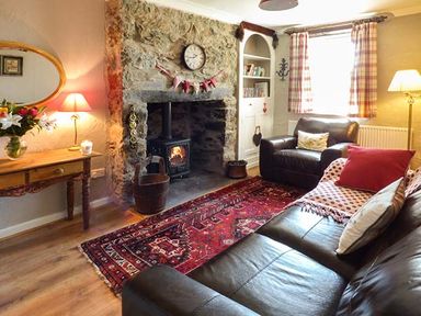 A living room with a stone fireplace and wood stove two leather sofas a wooden table with a lamp and flowers and a red patterned rug at Bwthyn Megan in Llangaffo near Newborough