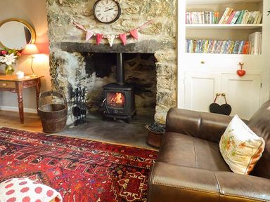 A living room with a stone fireplace wood burning stove a brown leather sofa and a bookcase filled with books and DVDs at Bwthyn Megan in Llangaffo near Newborough