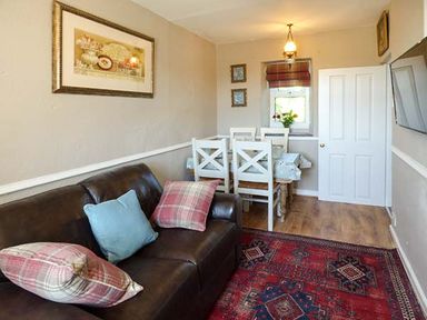 A seating area with a brown sofa and dining table with white chairs near a window at Bwthyn Megan in Llangaffo near Newborough
