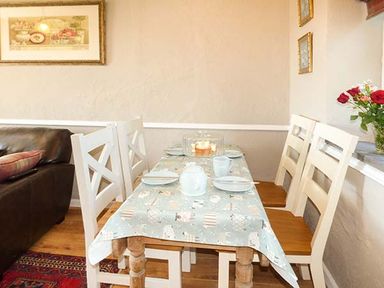 A dining table with four white chairs set with plates cups and a tablecloth next to a sofa with a cushion at Bwthyn Megan in Llangaffo near Newborough
