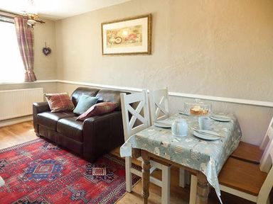 A room with a brown sofa with cushions a small dining table with four white chairs and a framed picture on the wall at Bwthyn Megan in Llangaffo near Newborough