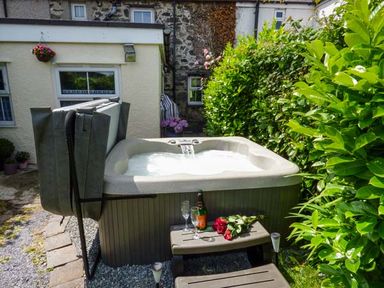 An outdoor hot tub with water flowing into it surrounded by green bushes and a small table with wine glasses and flowers at Bwthyn Megan in Llangaffo near Newborough