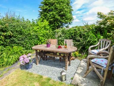 An outdoor seating area with a wooden table and chairs surrounded by plants and bushes at Bwthyn Megan in Llangaffo near Newborough