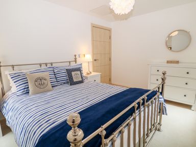 A bedroom with a metal bed frame and striped bedding a lamp on a nightstand and a dresser with a round mirror at The Creekside in Looe