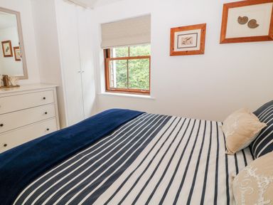 A bedroom with a striped bedspread two pillows a window dresser and framed pictures on the wall at The Creekside in Looe