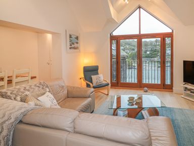 A living room with a leather sofa, glass coffee table, armchair, floor lamp, and large windows overlooking a hillside at The Creekside in Looe