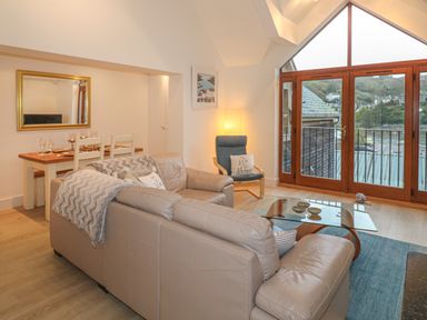 A living room with a beige leather sectional sofa a glass coffee table a dining table with chairs and large windows showing a view outside at The Creekside in Looe