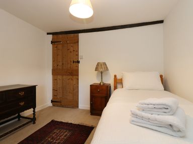 A bedroom with a single bed white bedding folded towels a wooden bedside table with a lamp a wooden dresser a wooden door and a rug on the floor at 13 Castle Hill in Lostwithiel