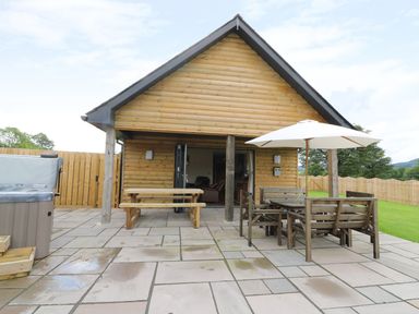 A wooden cabin with outdoor picnic tables umbrellas and a hot tub on a paved patio at Caban Nant Ddu in Rhayader