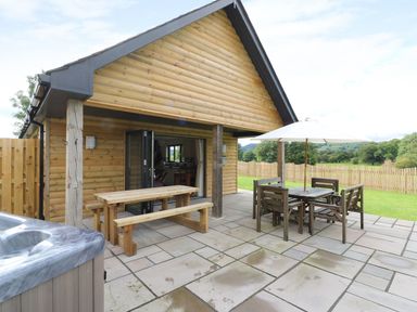 An outdoor patio area with a wooden picnic table bench set a dining table with an umbrella and hot tub at Caban Nant Ddu in Rhayader