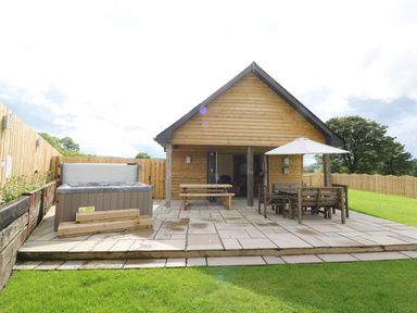 A wooden cabin with a patio featuring a hot tub and outdoor dining furniture with an umbrella at Caban Nant Ddu in Rhayader