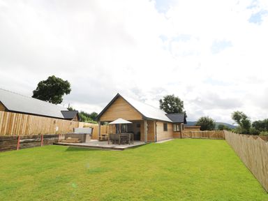 Wooden cabin with patio table and umbrella on a deck in a fenced yard with green grass at Caban Nant Ddu in Rhayader