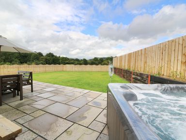 An outdoor patio with stone tiles a hot tub wooden table with chairs under a large umbrella and a grassy fenced yard at Caban Nant Ddu in Rhayader