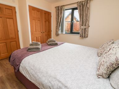 A bedroom with a bed made with patterned bedding and folded towels near wooden doors and a window with patterned curtains at Caban Nant Ddu in Rhayader