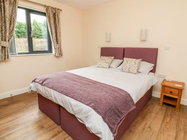 A bedroom with a double bed with purple headboard and throw beside a wooden nightstand with window and curtains at Caban Nant Ddu in Rhayader