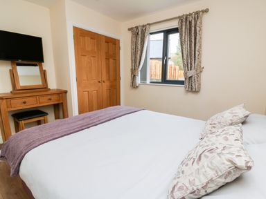 A bedroom with a bed with pillows and blanket a wooden dressing table with mirror stool a wall-mounted TV and a window with curtains at Caban Nant Ddu in Rhayader