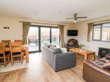 A living room with a gray sofa, wooden coffee table with flowers, brown leather chairs, dining table with chairs, ceiling fan, and sliding glass doors at Caban Nant Ddu in Rhayader