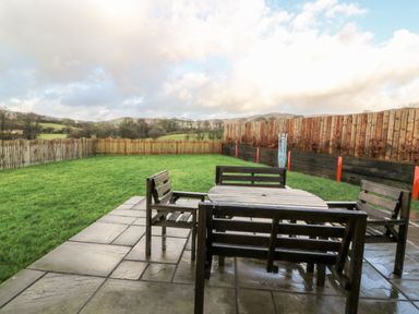 A patio with wooden table and chairs overlooking a fenced grassy yard and hills at Caban Nant Ddu in Rhayader