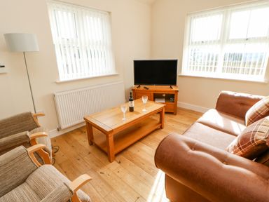 A living room with a brown sofa two armchairs a wooden coffee table with a bottle and two glasses a wooden TV stand and a television at Wansbeck Longframlington near Alnwick