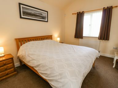 A bedroom with a wooden bed frame a white patterned bedspread two bedside tables with lamps a window with brown curtains and a framed picture above the bed at Wansbeck in Longframlington near Alnwick