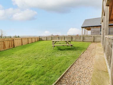 A fenced garden with green grass a wooden picnic table with bottles and glasses next to a wooden house at Wansbeck in Longframlington near Alnwick