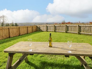 A wooden picnic table with a bottle and two wine glasses on a grassy fenced backyard at Wansbeck in Longframlington near Alnwick