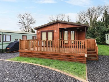A wooden cabin with a front porch and railing surrounded by gravel and grass at Roe Deer Lodge in Castlewigg near Whithorn