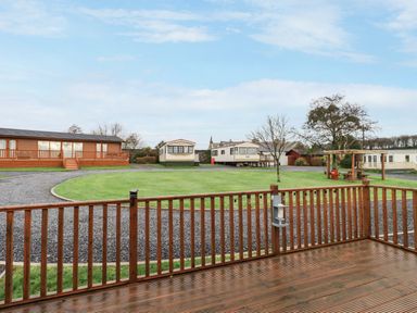 A wooden deck with railing overlooking a grassy area with gravel paths and several mobile homes at Roe Deer Lodge Castlewigg near Whithorn