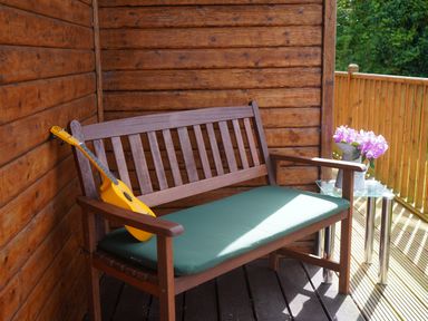 A wooden bench with a green cushion and a yellow guitar resting on it next to a glass table with flowers on a wooden covered porch at Roe Deer Lodge Castlewigg near Whithorn