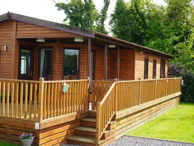 A wooden cabin with a deck and railing surrounded by grass and trees at Roe Deer Lodge Castlewigg near Whithorn
