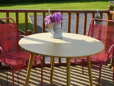A round table with a metal pitcher of purple flowers and two red chairs on a wooden deck at Roe Deer Lodge in Castlewigg near Whithorn