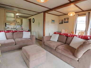 A living room with two brown sofas and a matching ottoman adjacent to an open kitchen and dining area at Roe Deer Lodge in Castlewigg near Whithorn