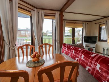 A dining area with wooden chairs and a bowl of apples on the table next to a living room with a sofa covered with a red blanket with white deer patterns at Roe Deer Lodge Castlewigg near Whithorn