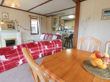 A dining table with a bowl of fruit and wooden chairs next to a living room with sofas and a kitchen at Roe Deer Lodge in Castlewigg near Whithorn