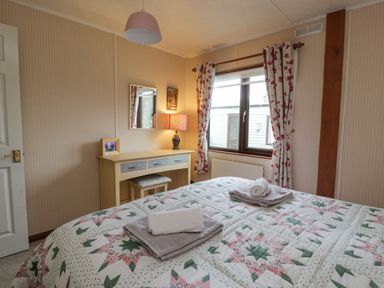 A bedroom with a bed covered in a patterned quilt and folded towels a desk with a mirror and lamp and a window with floral curtains at Roe Deer Lodge Castlewigg near Whithorn