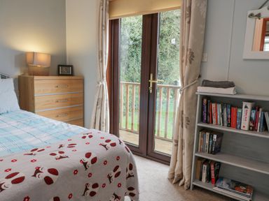A bedroom with a bed next to a wooden chest of drawers and a lamp and a glass door with curtains leading outside at Roe Deer Lodge Castlewigg near Whithorn