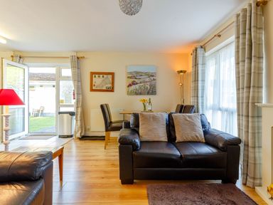 A living room with leather sofas a wooden dining table and chairs next to a glass door and large windows with curtains at Brook Cottage Rosecraddoc Lodge near Liskeard