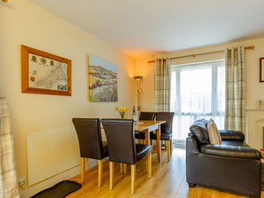 A dining area with a wooden table and four black chairs next to a window with plaid curtains and a black leather sofa in a living room at Brook Cottage Rosecraddoc Lodge near Liskeard